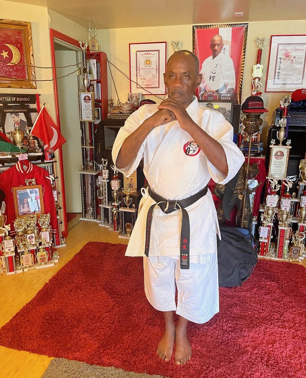 Shihan Abdur Rahiem in a white gi and black belt, standing in front of five decades of trophies and diplomas